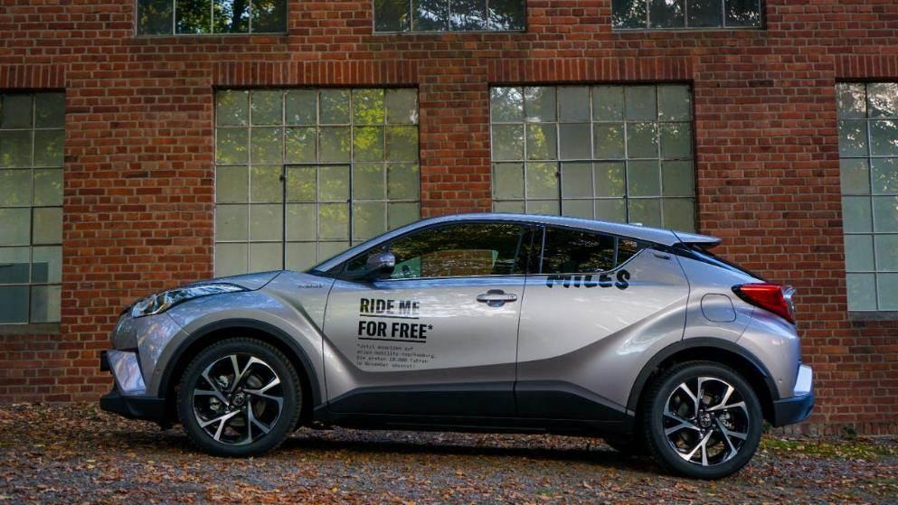 grauer Toyota CH-R in der Seitenansicht vor Fabrikgebäude mit der Aufschrift "Ride me for free"
